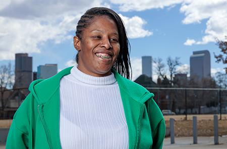 Tasha Clavo stands in front of the Denver skyline wearing a white turtleneck and kelly green zip up
